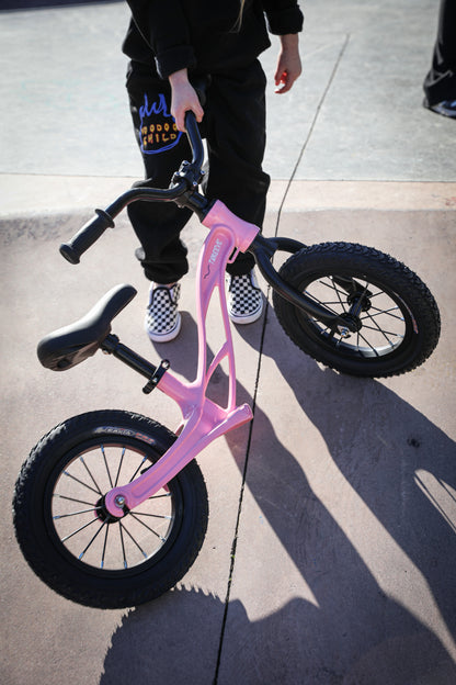 Pink balance bike at a skatepark, handlebars being held by a child in a black tracksuit