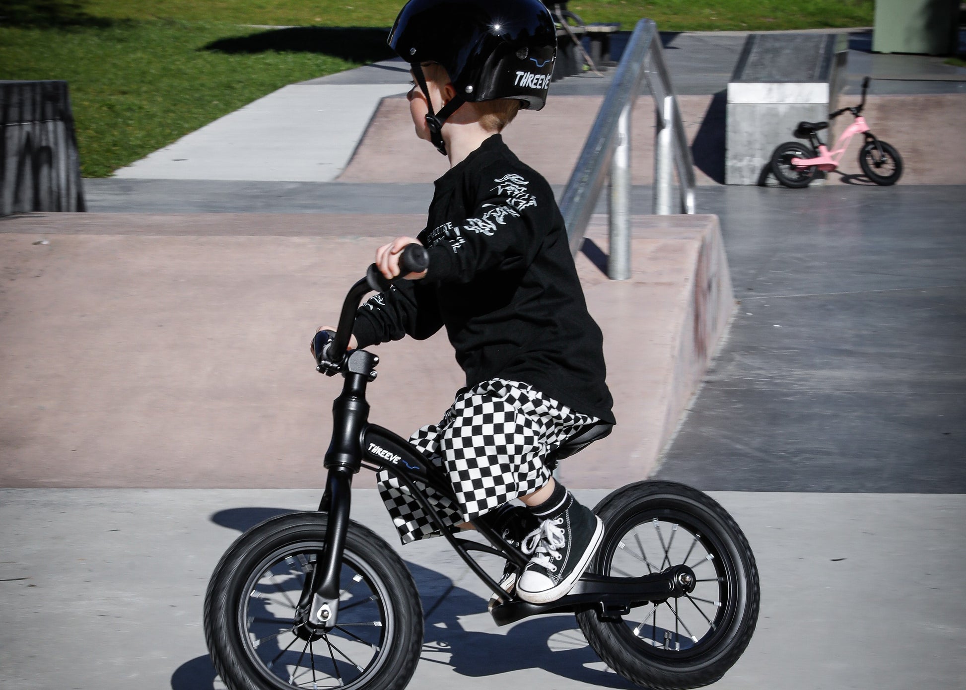 Toddler riding black threeve balance bike at skatepark, wearing black helmet