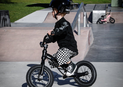 Toddler riding black threeve balance bike at skatepark, wearing black helmet